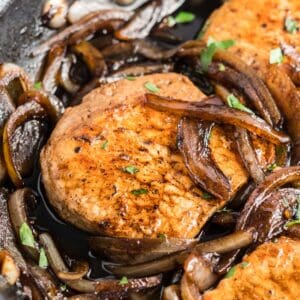 A close-up of seared pork chops with caramelized onions in a skillet, garnished with chopped parsley.