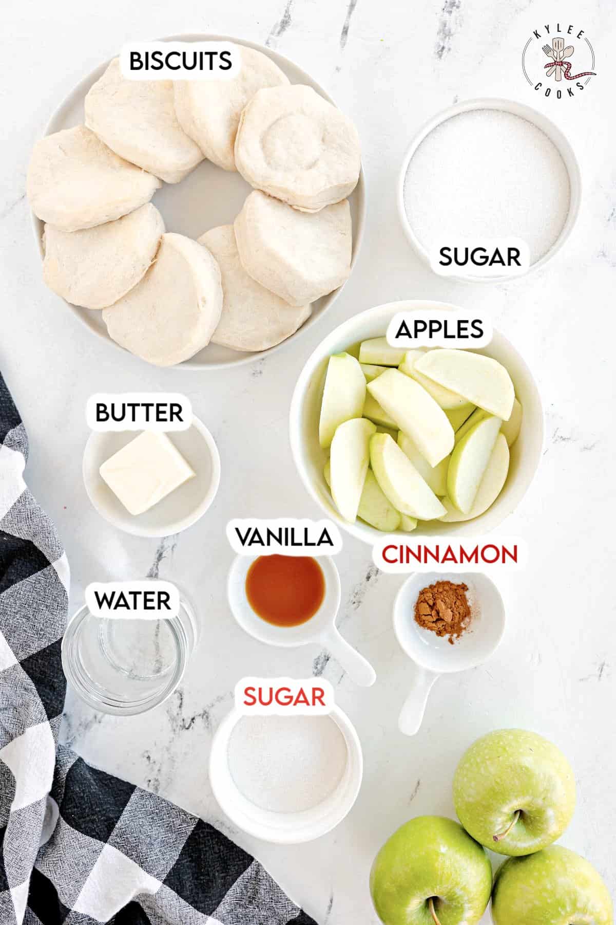 A flat lay of ingredients for a recipe, including biscuits, butter, water, vanilla, cinnamon, two bowls of sugar, a plate of apple slices, and three whole green apples.