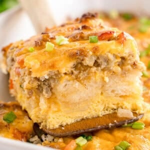 A slice of biscuit breakfast bake with melted cheese and green onions being lifted on a wooden spatula.