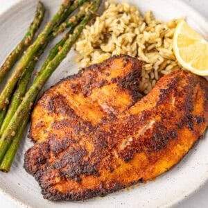 Blackened tilapia on a plate with rice, asparagus, and a lemon wedge.