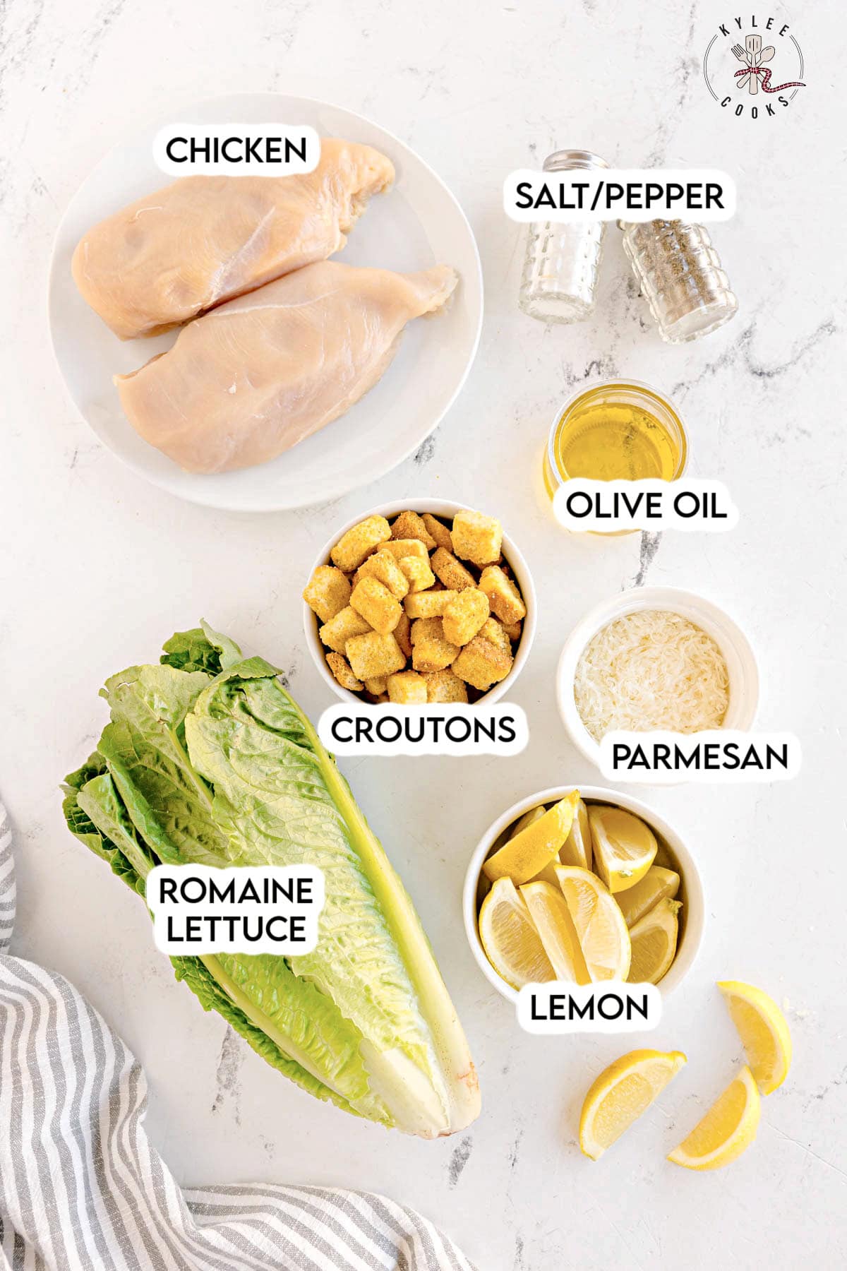 Raw chicken breasts on a plate, with bowls of croutons, parmesan, lemon wedges, and labeled ingredients including romaine lettuce, olive oil, and salt/pepper. Text on the image says 'CHICKEN', 'CROUTONS', 'PARMESAN', 'LEMON', 'ROMAINE LETTUCE', 'OLIVE OIL', 'SALT/PEPPER'.