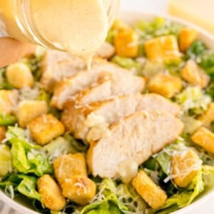 Sliced chicken on romaine lettuce with croutons and grated cheese, dressing being poured from a jar.