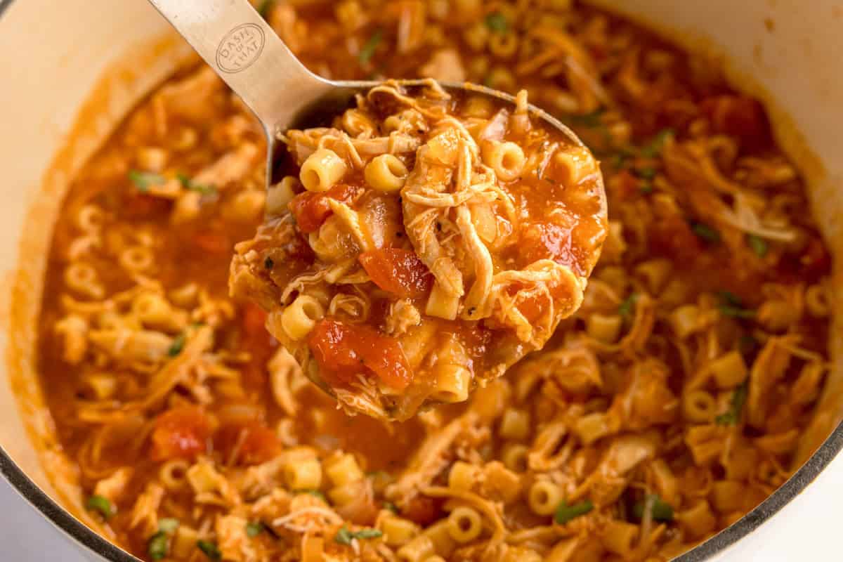 A ladle filled with chicken parmesan soup held above a pot, with visible tomatoes, pasta, and shredded chicken.
