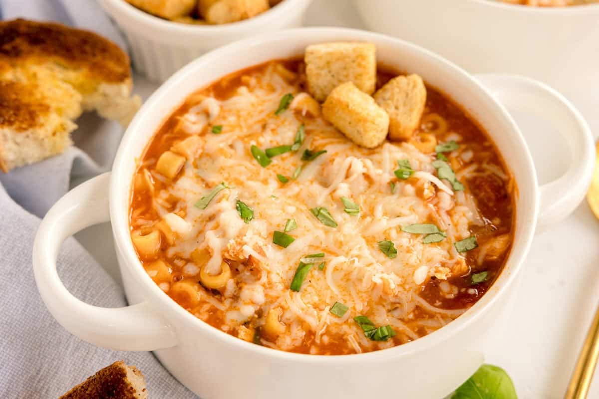 A white bowl of chicken parmesan soup topped with melted cheese, fresh basil, and croutons.