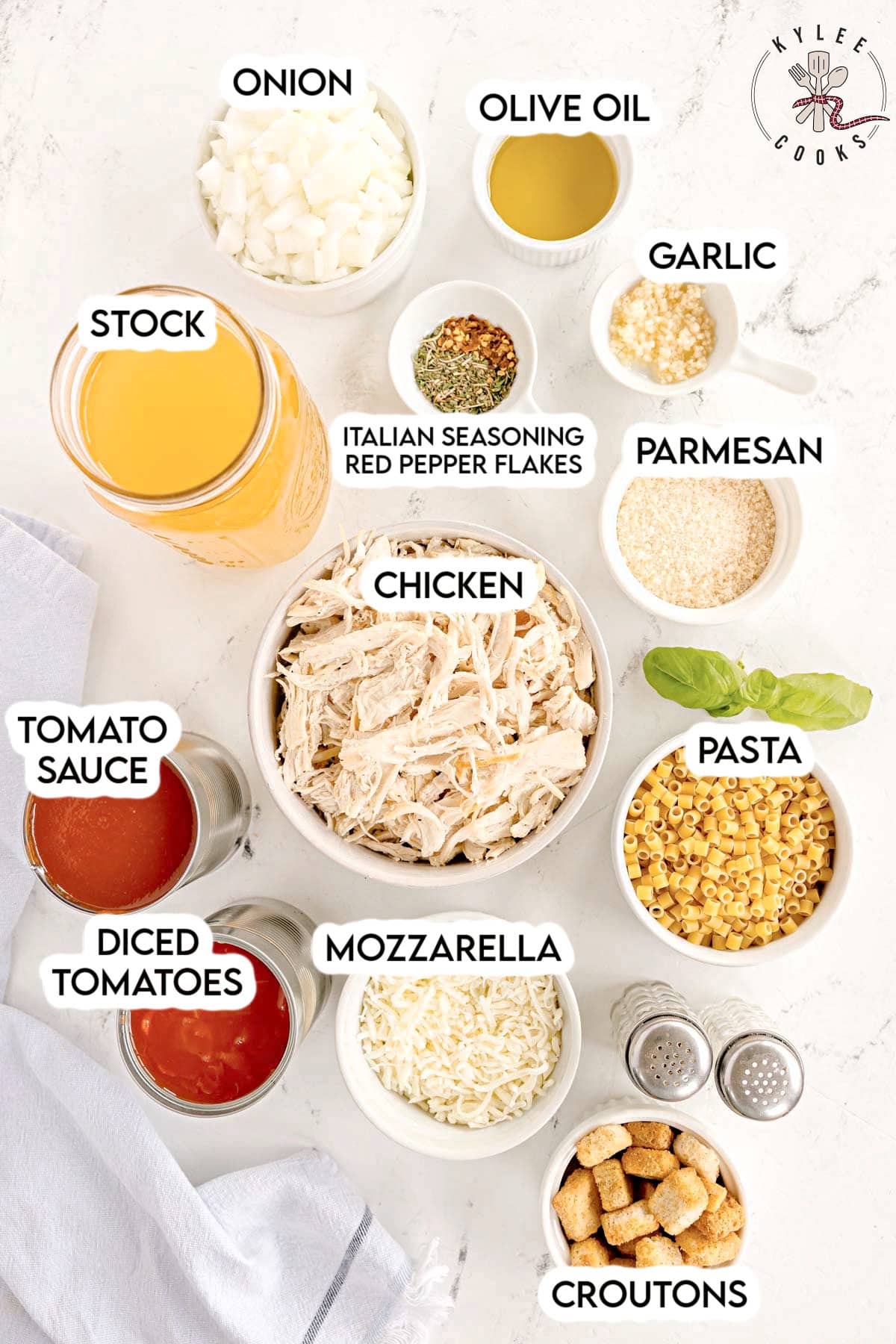 Bowls of chopped onion, olive oil, garlic, shredded chicken, pasta, parmesan, mozzarella, croutons, seasonings, tomato sauce, diced tomatoes, and stock. Text on the image says 'ONION', 'OLIVE OIL', 'GARLIC', 'CHICKEN', 'PASTA', 'PARMESAN', 'MOZZARELLA', 'CROUTONS', 'ITALIAN SEASONING RED PEPPER FLAKES', 'TOMATO SAUCE', 'DICED TOMATOES', 'STOCK'.