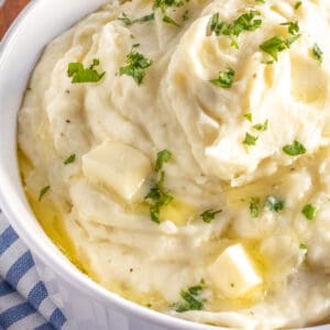 A bowl of mashed potatoes topped with melted butter and chopped herbs, placed on a striped cloth.