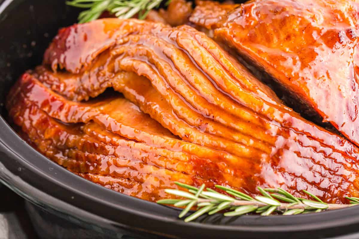 Sliced, glazed ham in a crock pot, garnished with fresh rosemary sprigs.