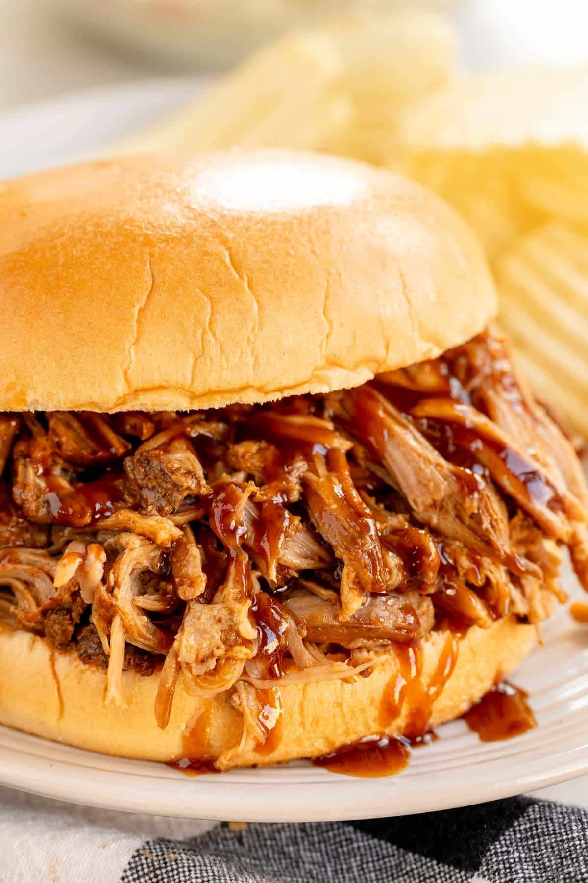 A pulled pork sandwich drenched in barbecue sauce on a white plate, accompanied by ridged potato chips.