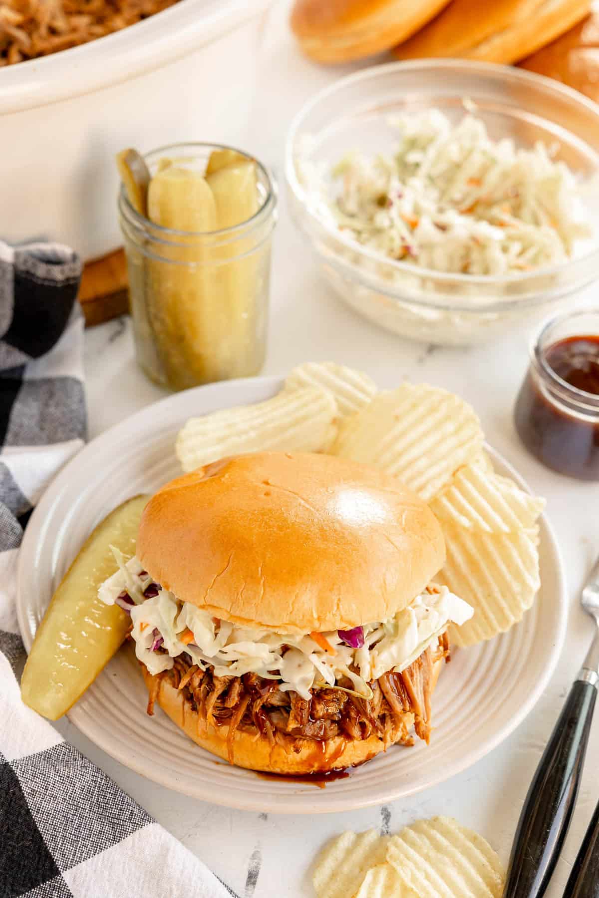 A plate with a pulled pork sandwich topped with coleslaw, surrounded by potato chips and a pickle. Nearby are a jar of pickles, a bowl of coleslaw, a cup of sauce, and a black and white checkered napkin.