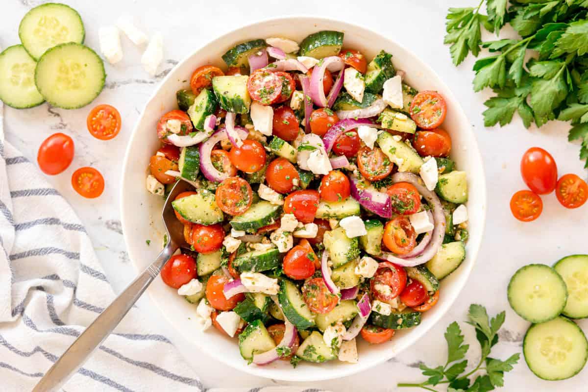 Overhead view of a bowl filled with cucumber salad, surrounded by herbs and veggies.