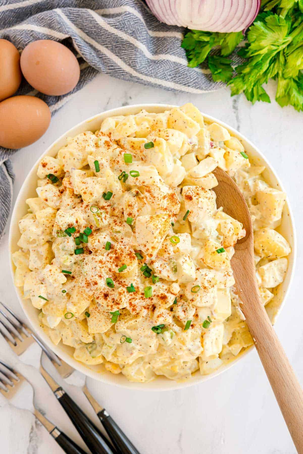 Overhead view of potato salad in a bowl with a wooden spoon, surrounded by eggs, celery, and red onion.