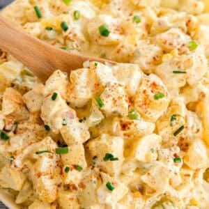 A wooden spoon scooping creamy potato salad topped with chopped chives and paprika.