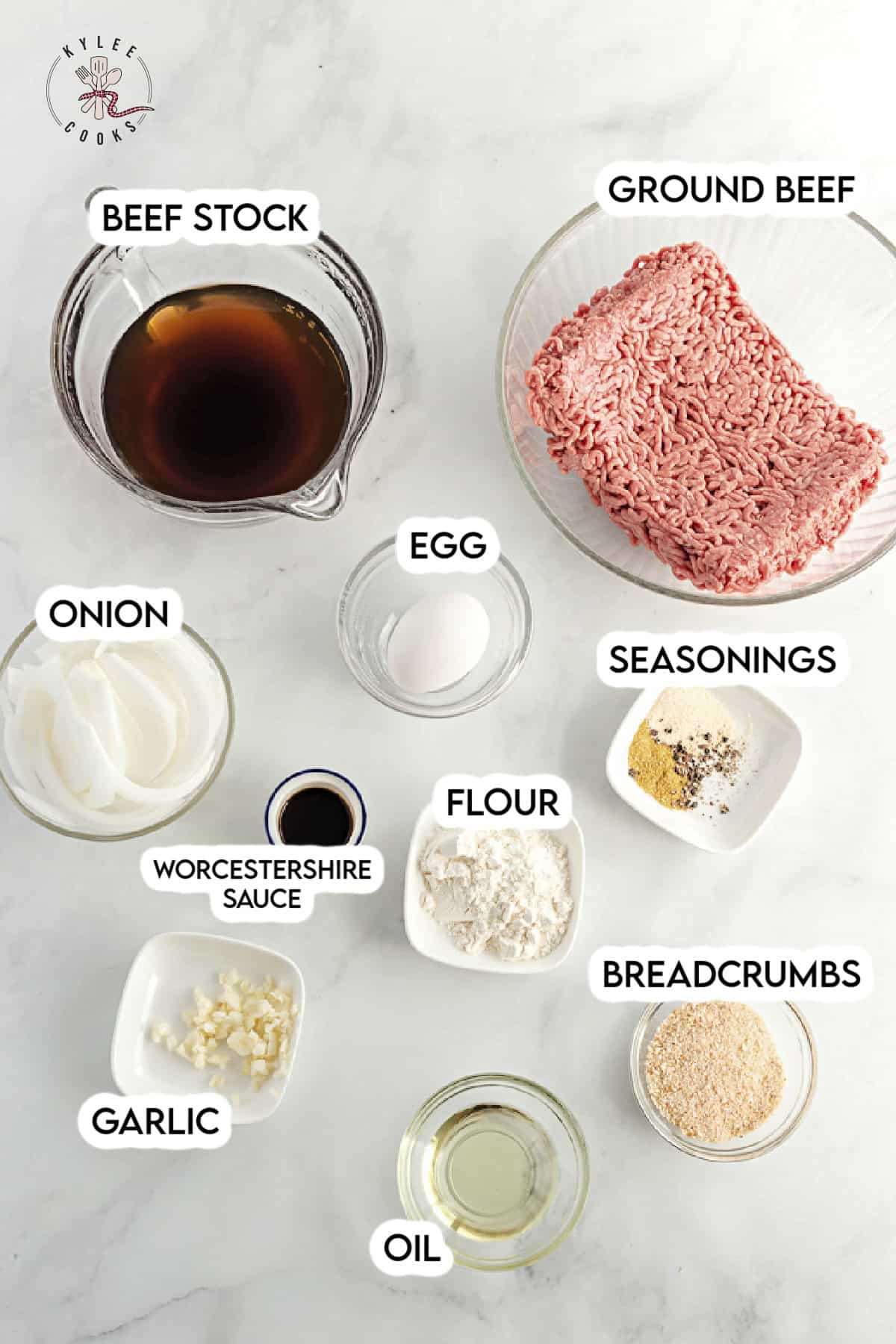 Overhead shot of ingredients for hamburger steaks with onion gravy, including ground beef, breadcrumbs, egg, seasonings, onions, flour, garlic, oil, Worcestershire sauce, and beef stock, each labeled.
