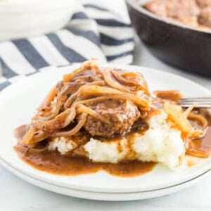 A serving of mashed potatoes topped with a hamburger steak, sautéed onions, and rich hamburger steaks with onion gravy on a white plate.