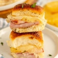 Two stacked ham and cheese sliders on soft rolls, topped with poppy seeds and garnished with chopped herbs, served on a white plate.