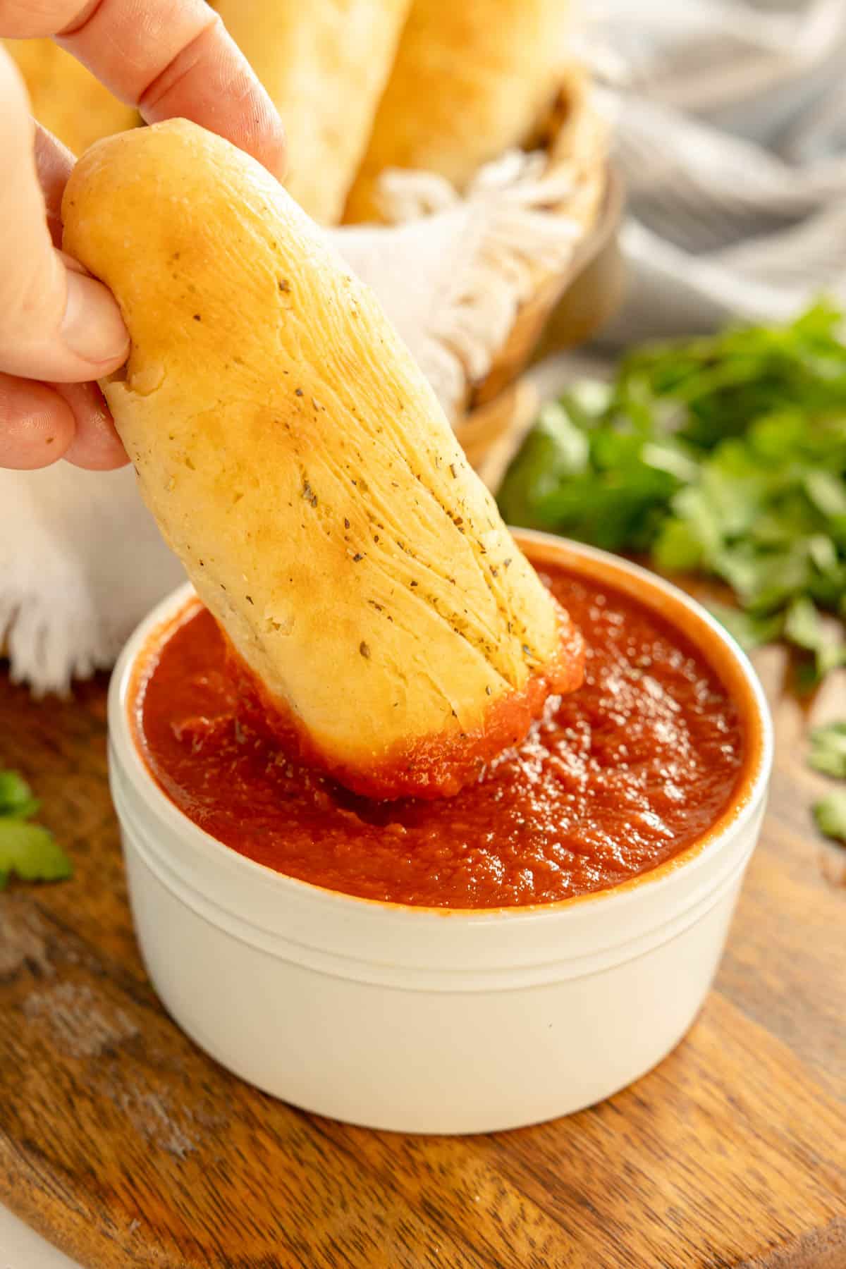 A hand dips a breadstick into a bowl of marinara sauce. A basket of more breadsticks is in the background, along with fresh parsley on a wooden surface.