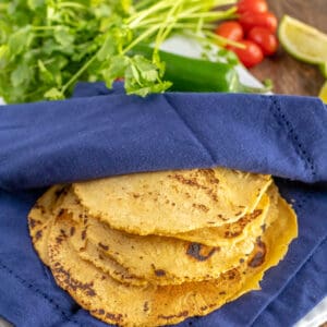 A stack of corn tortillas partially covered by a blue cloth napkin, with cilantro, tomatoes, lime wedges, and a jalapeño in the background.