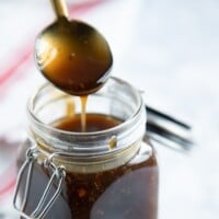 A spoon drizzling caramel-colored homemade teriyaki sauce into an open glass jar with a metal clasp, against a blurred background.