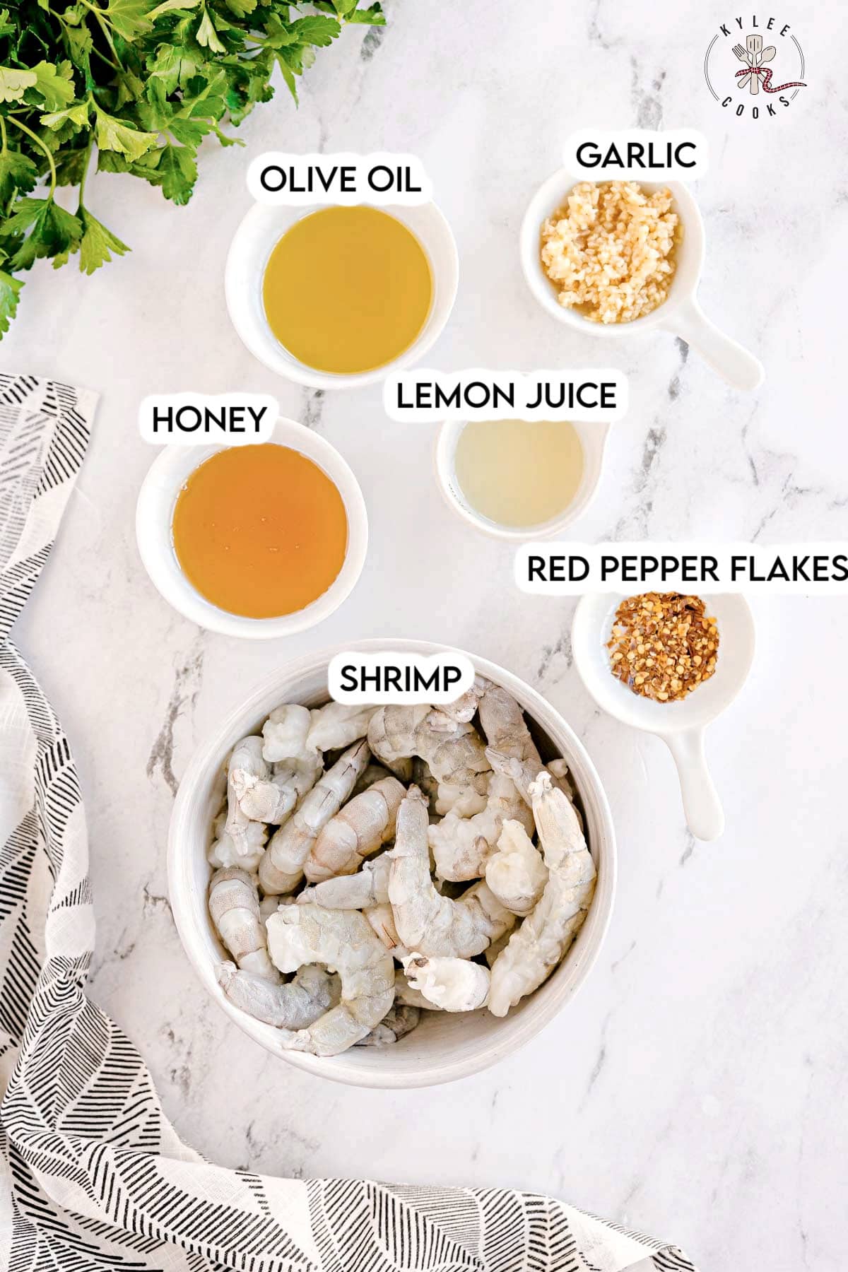 A bowl of raw shrimp surrounded by small bowls of olive oil, garlic, honey, lemon juice, and red pepper flakes. Each bowl is labeled.
