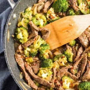 A close-up of a skillet filled with honey soy beef and broccoli, garnished with red pepper flakes, with a wooden spatula stirring the mixture.