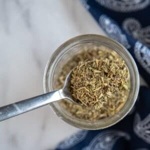 A spoon holding dried Italian seasoning blend is positioned over an open glass jar on a marble surface.