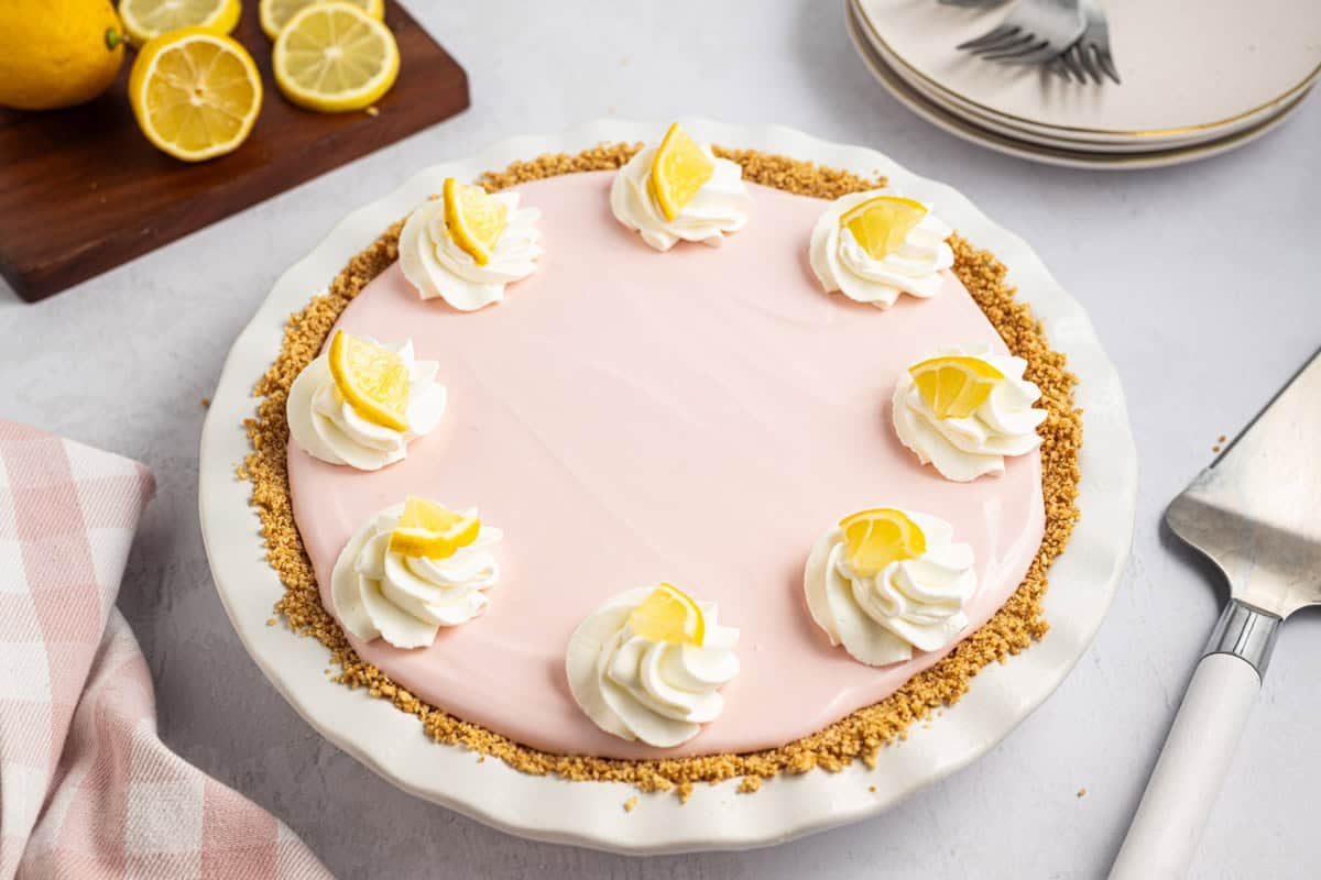 A pink lemonade pie in a white pie dish with a graham cracker crust and swirls of whipped cream topped with lemon slices around the edge. Plates, a cake server, and lemons are nearby.