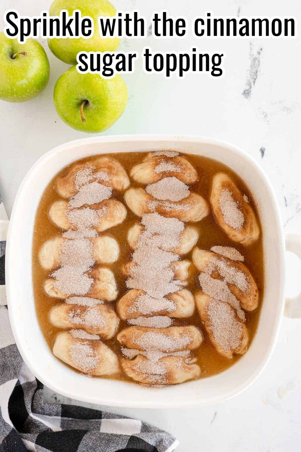 A white baking dish filled with dough pieces covered in a cinnamon sugar topping and surrounded by a brown liquid. Green apples and a checkered cloth are placed nearby.