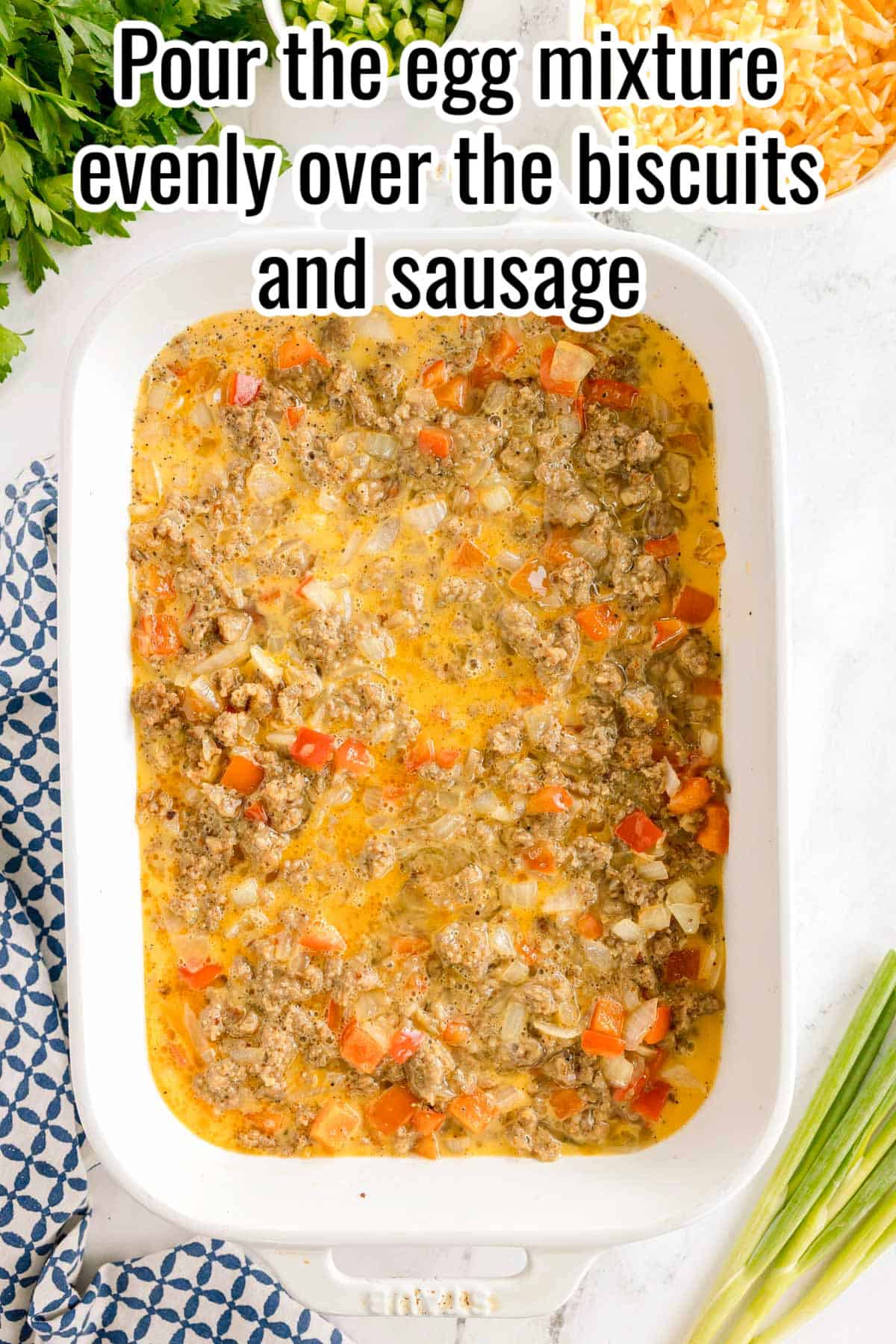 Egg mixture poured over sausage and biscuit layers in a baking dish. Text on the image says 'Pour the egg mixture evenly over the biscuits and sausage'.