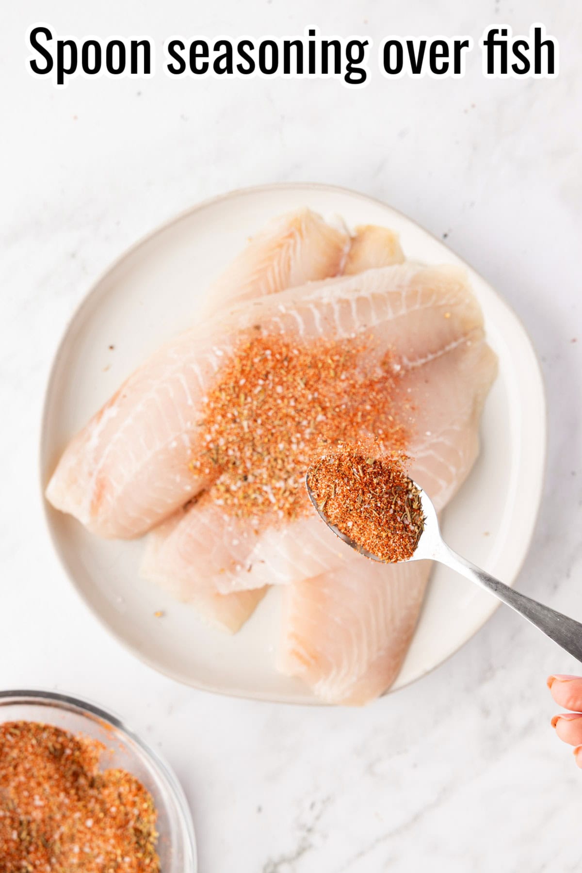 Spoonful of seasoning being sprinkled over raw tilapia fillets. Text on the image says 'Spoon seasoning over fish'.