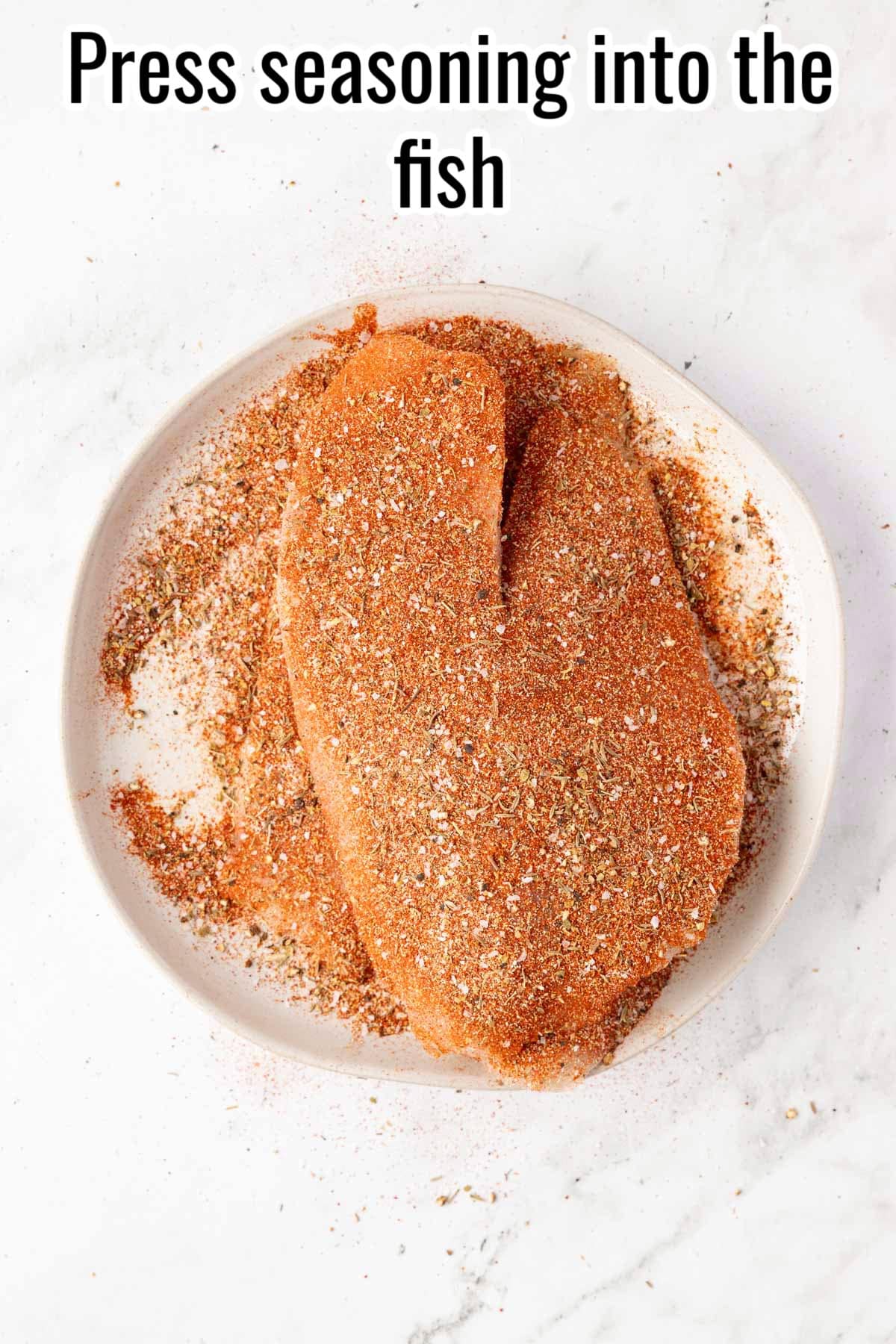Raw tilapia fillets fully coated in seasoning. Text on the image says 'Press seasoning into the fish'.