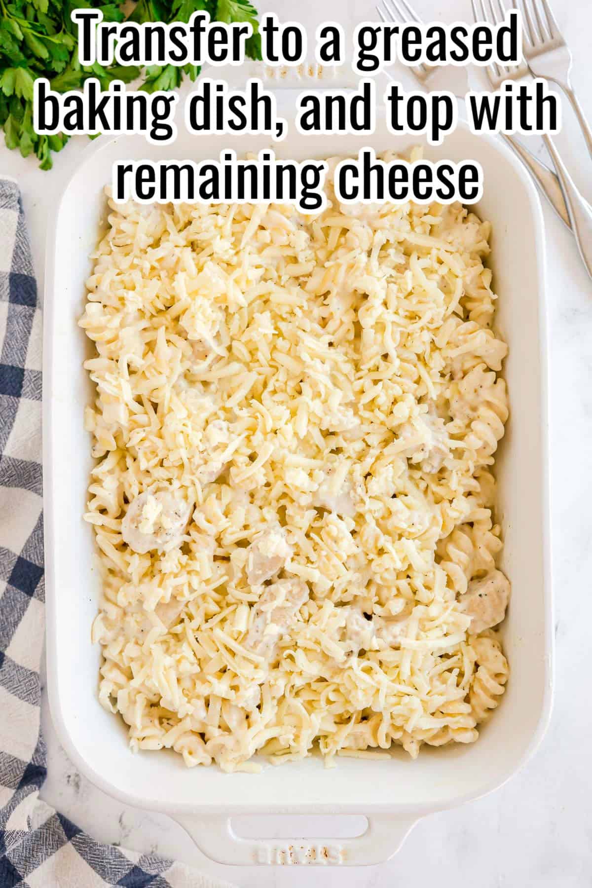 Pasta mixture in a white baking dish topped with shredded cheese. Text on the image says 'Transfer to a greased baking dish, and top with remaining cheese'.