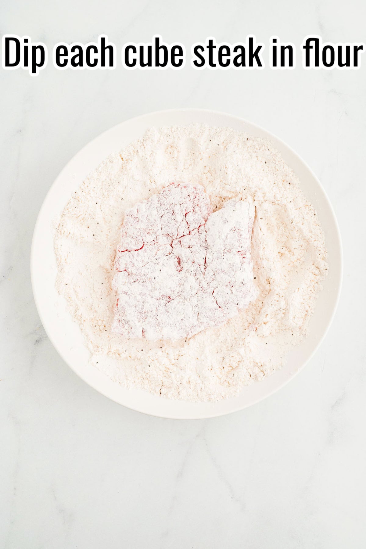 Cube steak coated in flour in a shallow bowl. Text on the image says 'Dip each cube steak in flour'.
