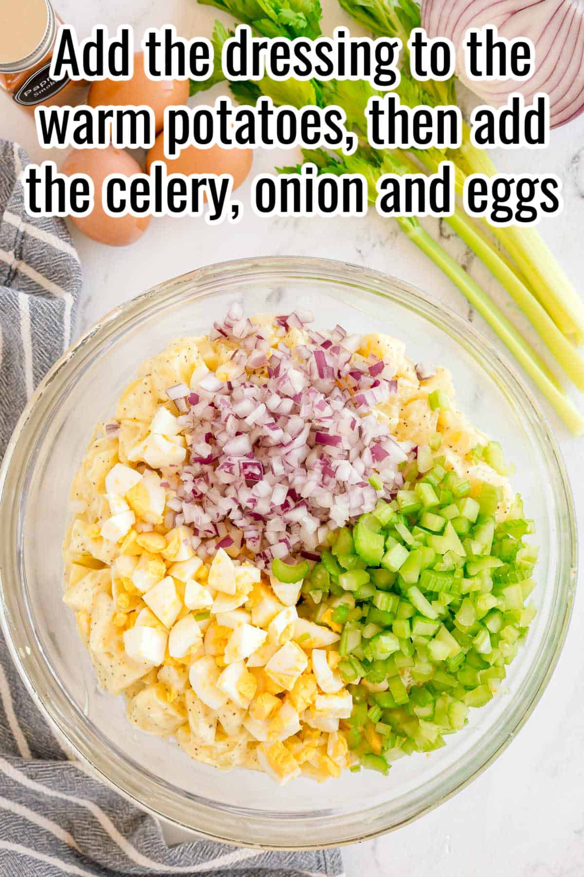 A large bowl with chopped celery, onion, and eggs on top of potato salad. Text on the image says 'Add the dressing to the warm potatoes, then add the celery, onion and eggs'.