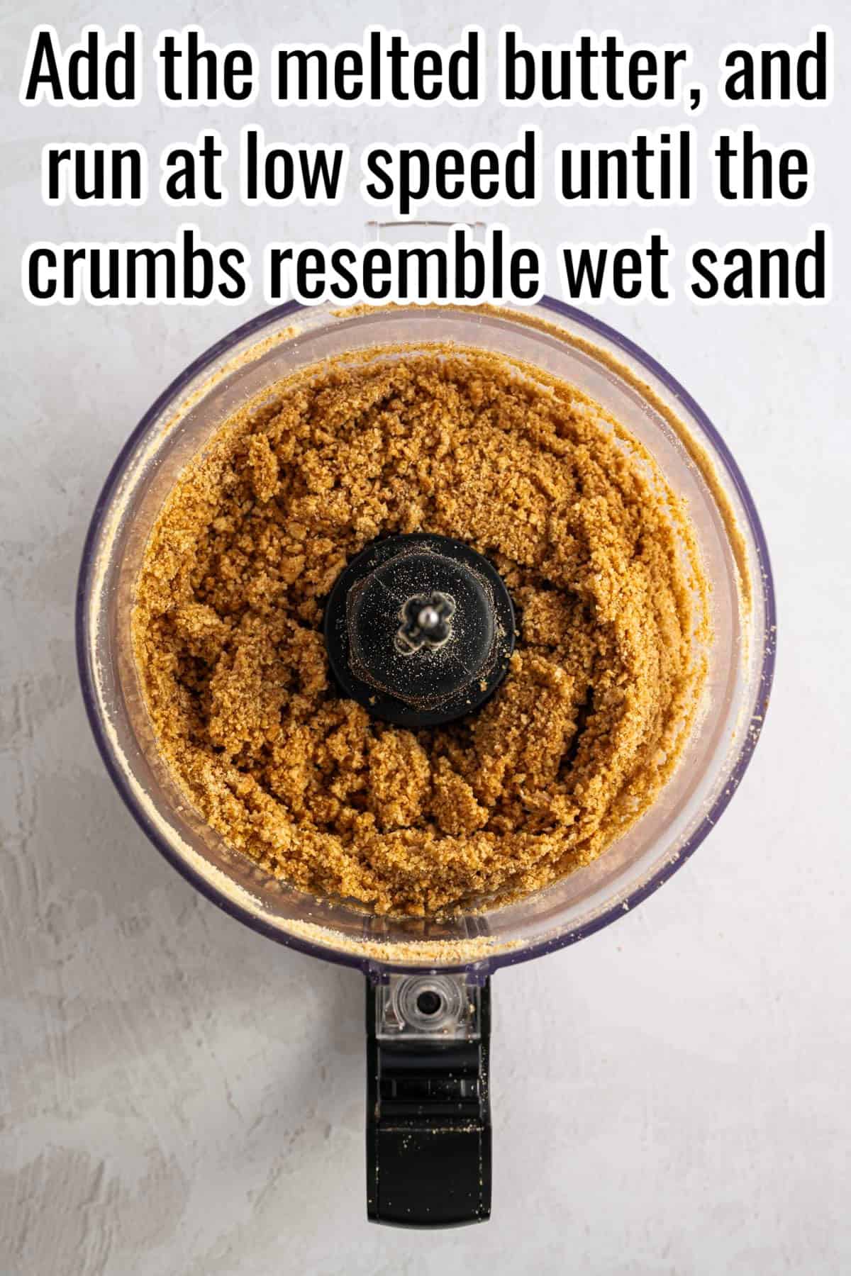 Crushed graham cracker crumbs in a food processor. Text on the image says 'Add the melted butter, and run at low speed until the crumbs resemble wet sand'.