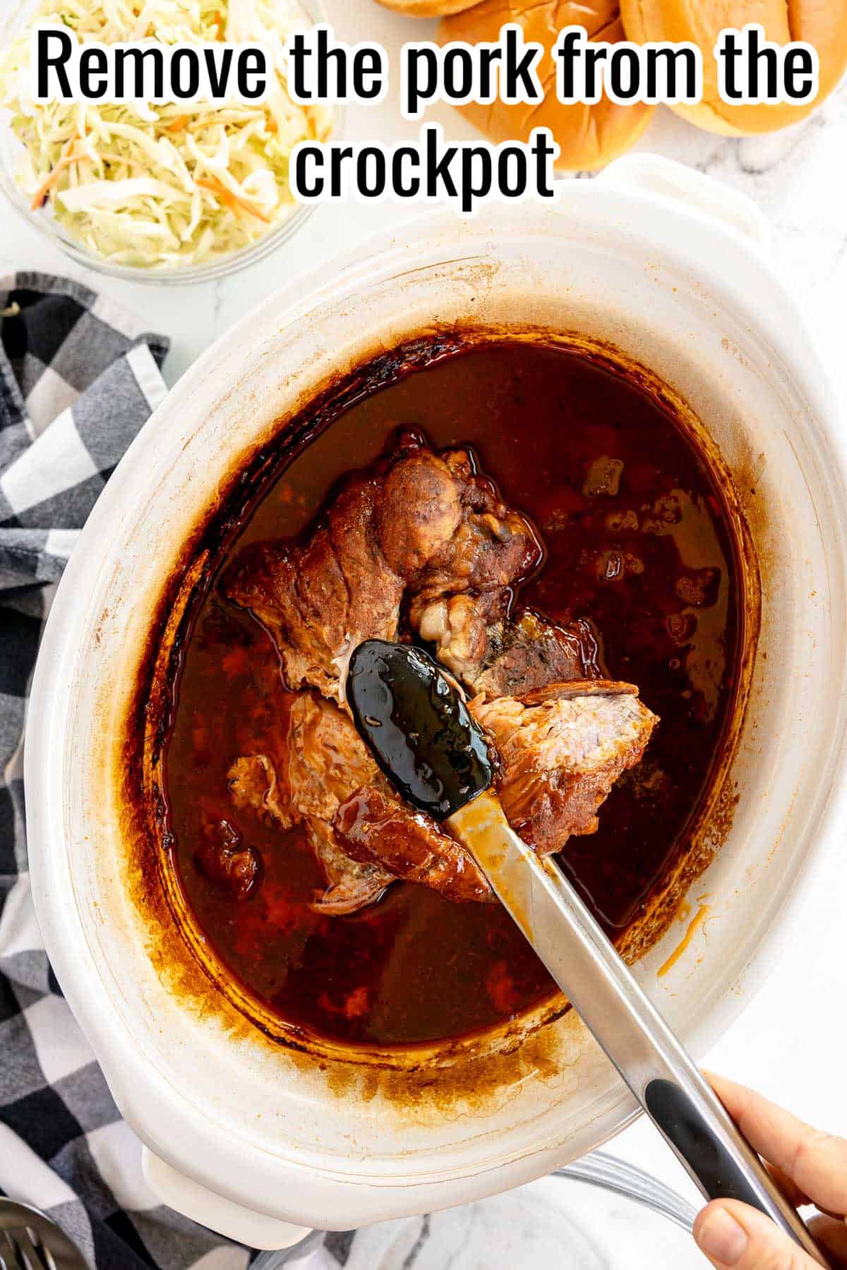 Hand holding tongs removing cooked pork from a crockpot filled with sauce. A bowl of coleslaw and a checkered cloth are visible in the background. Text reads: "Remove the pork from the crockpot.
