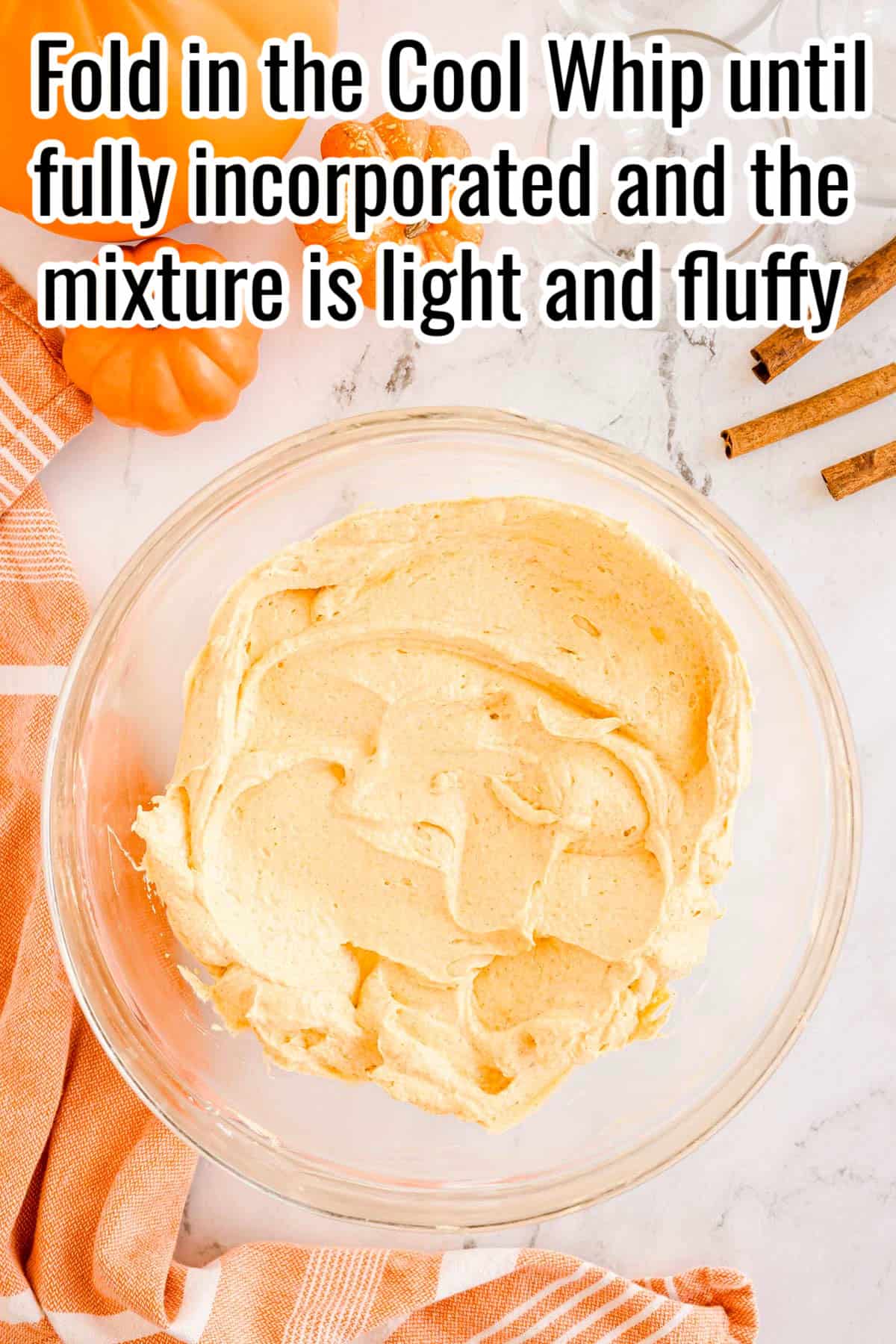 A glass bowl with Cool Whip folded into the pumpkin mixture until light and fluffy. Text on the image says 'Fold in the Cool Whip until fully incorporated and the mixture is light and fluffy'.