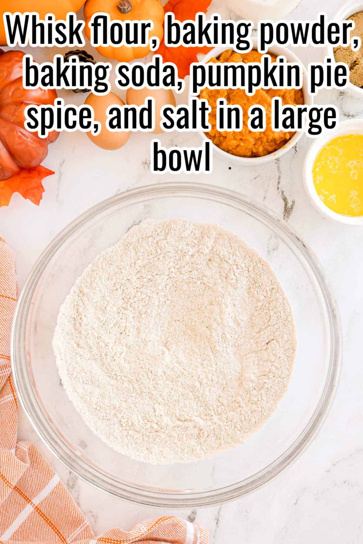 A glass bowl filled with flour mixture. Text on the image says 'Whisk flour, baking powder, baking soda, pumpkin pie spice, and salt in a large bowl'.