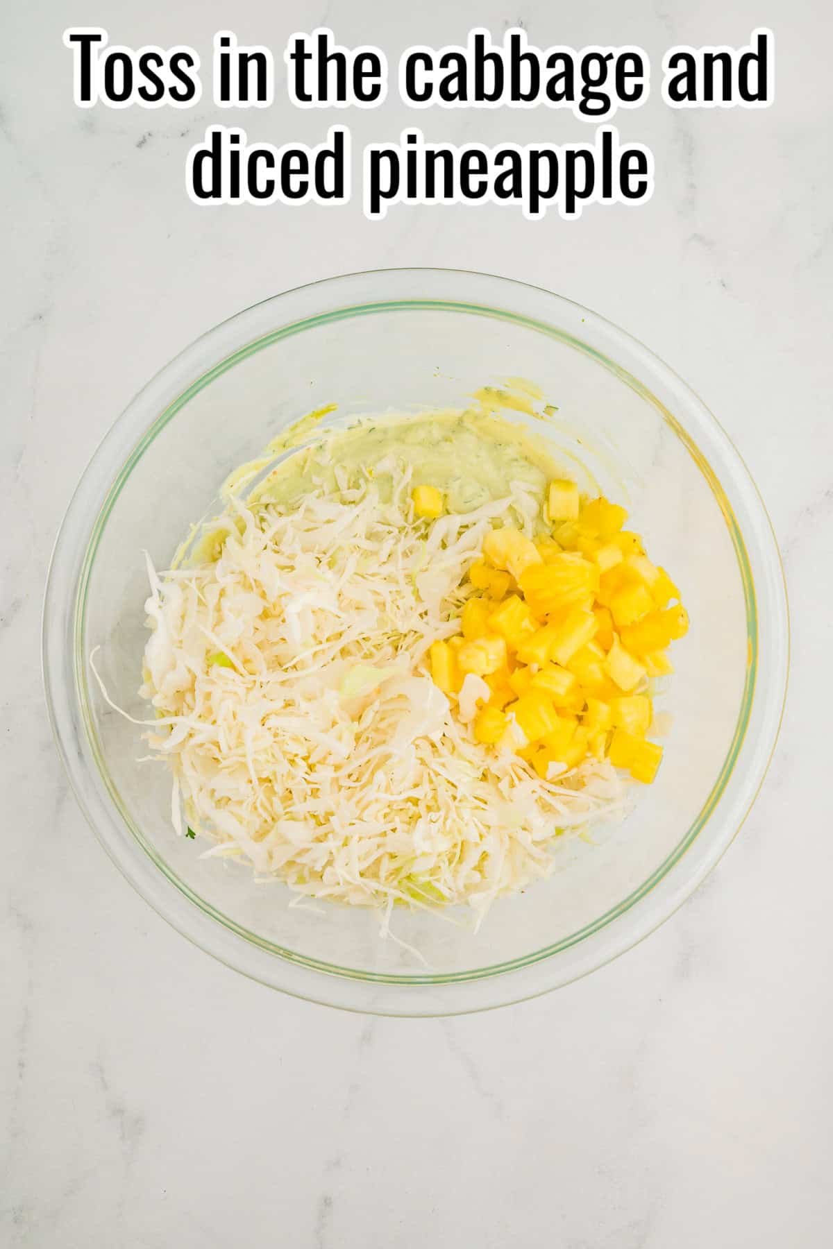 Shredded cabbage and diced pineapple on top of avocado dressing in a glass bowl. Text on the image says 'Toss in the cabbage and diced pineapple'.