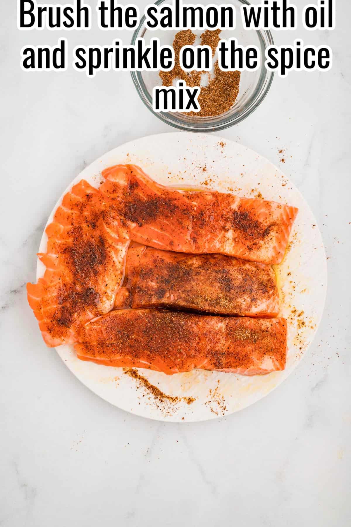 Raw salmon fillets brushed with oil and sprinkled with spice mix on a white plate. Text on the image says 'Brush the salmon with oil and sprinkle on the spice mix'.