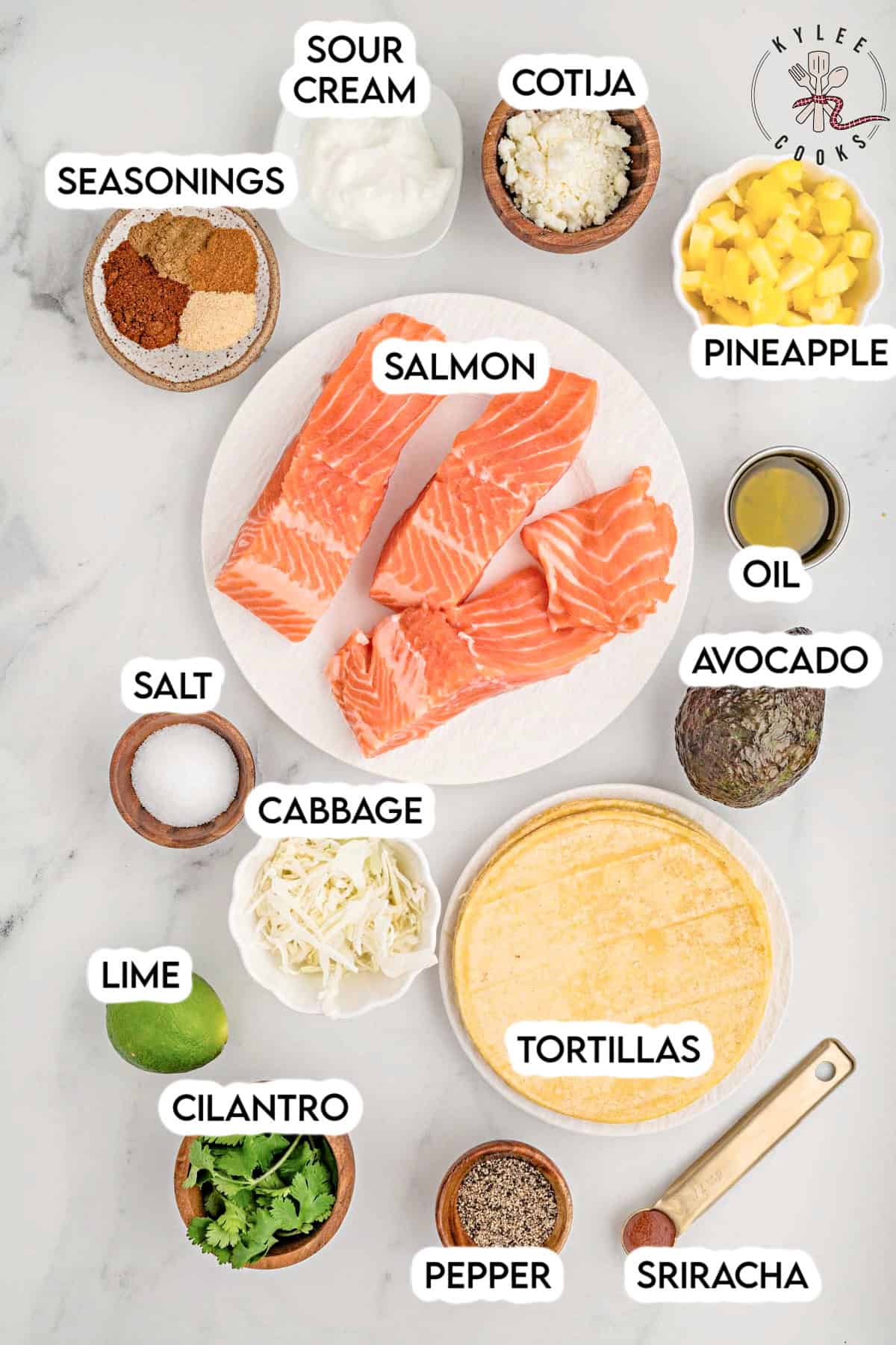 Ingredients for salmon tacos laid out, each labeled: salmon, sour cream, cotija, pineapple, oil, avocado, tortillas, pepper, sriracha, cilantro, lime, cabbage, salt, seasonings.