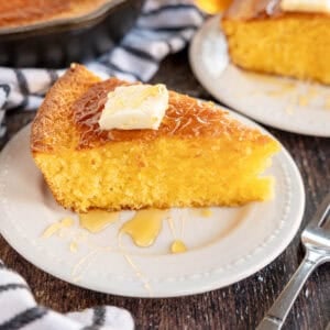 A slice of cornbread topped with a pat of butter and drizzled with honey sits on a white plate next to a fork.