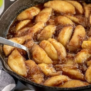 Sliced apples coated in a brown sugar sauce are being cooked in a cast iron skillet, with a metal spoon resting in the dish.