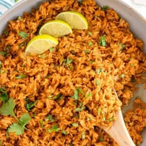 A pan of Spanish rice with lime wedges and cilantro, stirred with a wooden spoon.