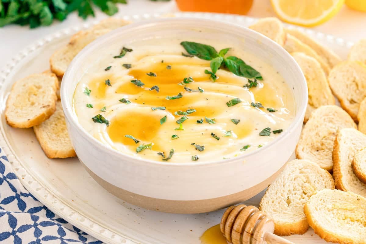 A bowl of creamy dip garnished with herbs and honey, surrounded by slices of toasted baguette on a white plate.