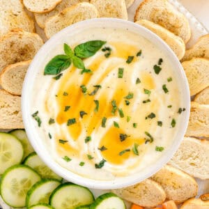 Overhead view of whipped ricotta dip topped with honey and herbs, surrounded by toasted bread slices and cucumber slices.